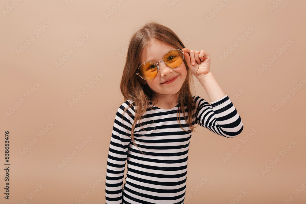 Happy smiling cute little girl with wearing orange round glasses and striped dress touching glasses and looking at camera over beige background Happy smiling cute little girl with wearing orange round glasses and striped dress touching glasses and looking at camera over beige background