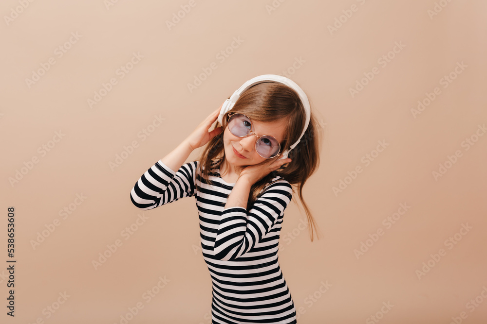 Joyful good-humored lovely little girl in striped dress rejoices and rises arms on isolated beige background. Charming happy girl listening music and having fun Joyful good-humored lovely little girl in striped dress rejoices and rises arms on isolated beige background. Charming happy girl listening music and having fun