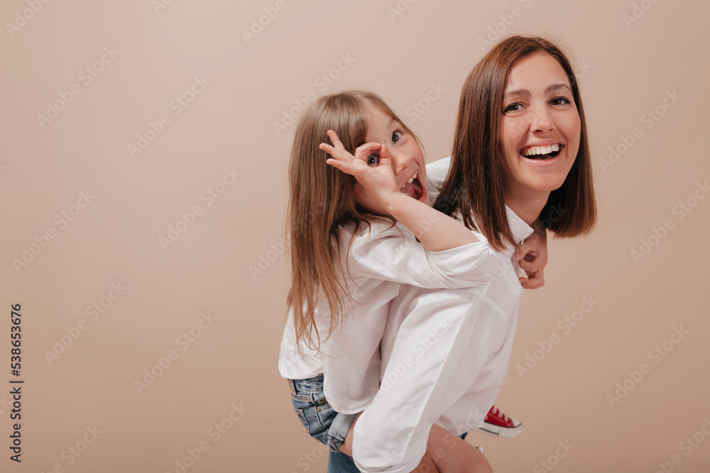 Happy excited little girl with mom having fun and smiling at camera over beige background. Adorable little girl shows ok sign and playing with mother