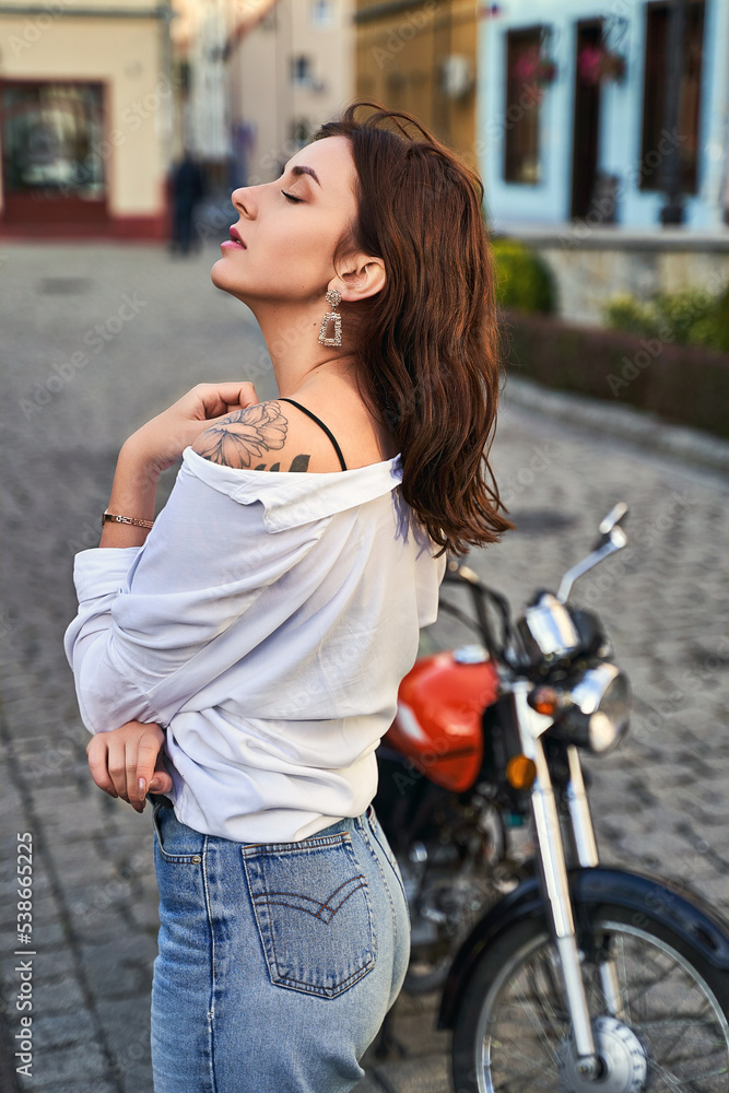 Young beautiful woman shows her bare shoulder with a tattoo standing by a bike on the street