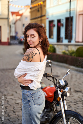 Obraz na płótnie Young beautiful woman shows her bare shoulder with a tattoo standing by a bike on the street w salonie