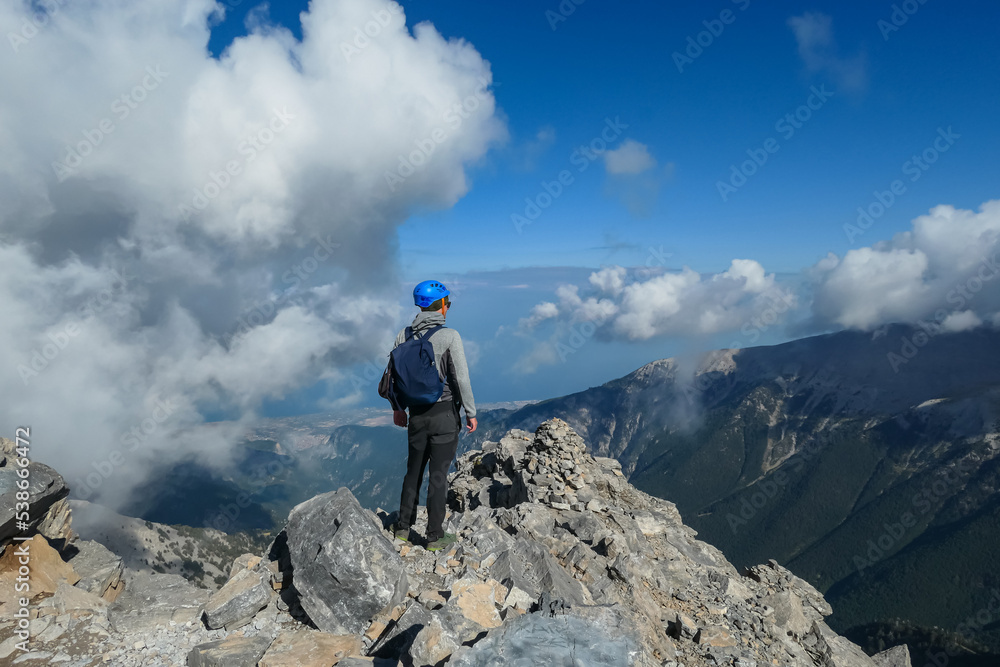 Rear view of man with climbing helmet on cloud covered mountain summit of Mytikas Mount Olympus, Mt Olympus National Park, Macedonia, Greece, Europe. View of rocky ridges and Mediterranean Aegean Sea
