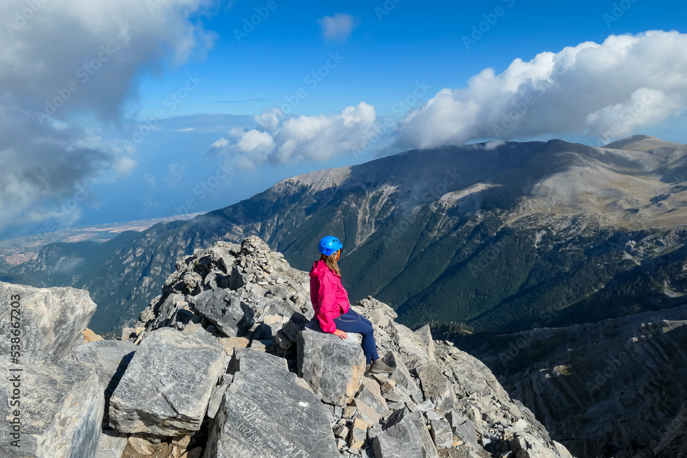 Rear view of woman with climbing helmet on cloud covered mountain summit Mytikas Mount Olympus, Mt Olympus National Park, Macedonia, Greece, Europe. View of rocky ridges and Mediterranean Aegean Sea