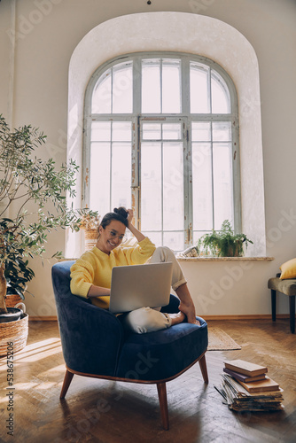 Attractive young woman using laptop and smiling while sitting in a comfortable chair at home