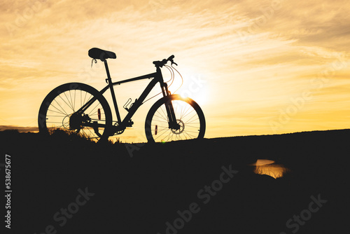 Fototapeta Naklejka Na Ścianę i Meble -  bike silhouette on mountain near river, orange sunset sky and blurry clouds
