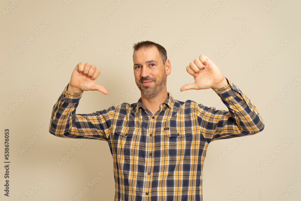 Studio portrait of funny proud man in his 40s pointing thumbs to himself, saying he's really cool, looking successful and confident, wearing shirt, standing over beige background, with copy space. Studio portrait of funny proud man in his 40s pointing thumbs to himself, saying he's really cool, looking successful and confident, wearing shirt, standing over beige background, with copy space.