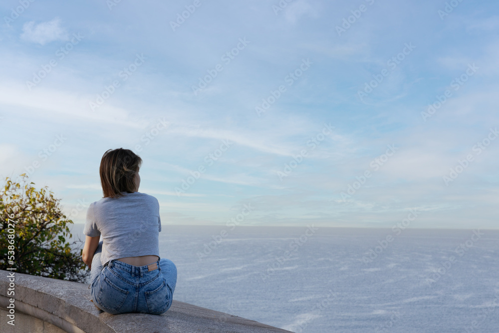 A young woman in jeans and a T-shirt sits on a stone slab and looks at the sea. High quality photo
