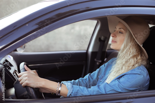 Young woman with long fair hair in denim shirt, beige floppy hat, sitting in black car, holding steering wheel, driving. © Татьяна Волкова