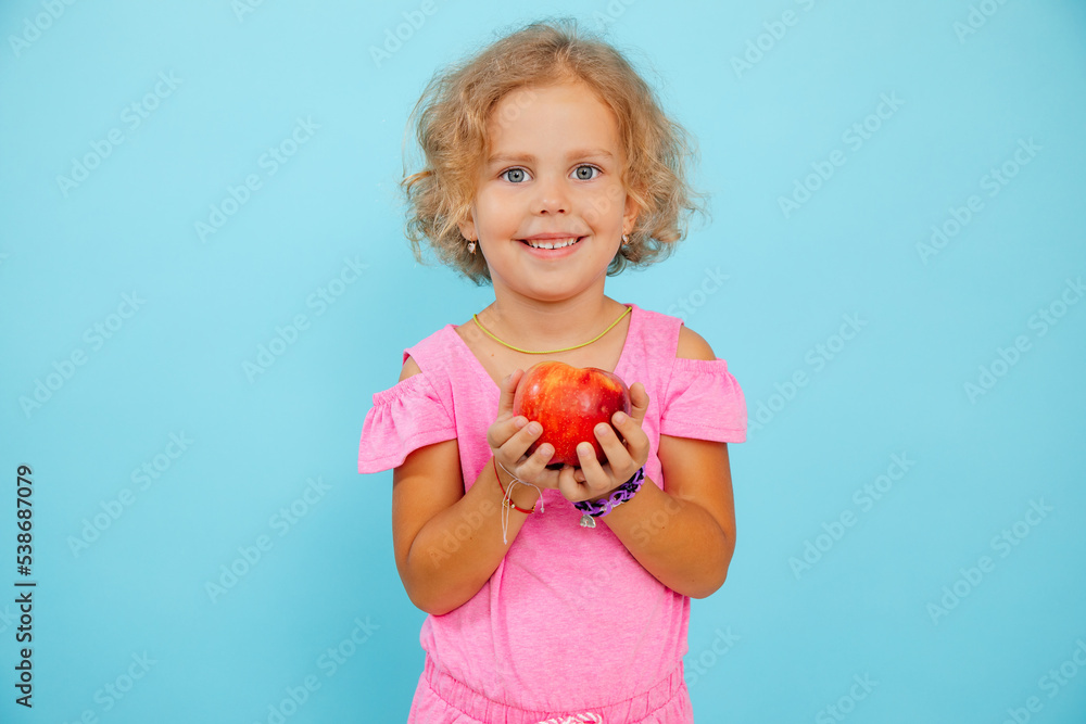 Small little smiling, expressive, positive curly blonde girl holding red juice apple in hands on blue view. Healthy life Small little smiling, expressive, positive curly blonde girl holding red juice apple in hands on blue view. Healthy life