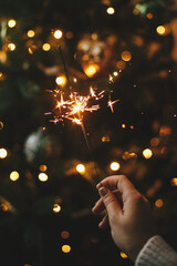 Happy New Year! Burning sparkler in female hand on background of christmas tree lights in dark room. Atmospheric celebration. Hand holding firework against stylish decorated tree with illumination