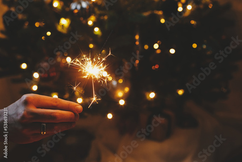 Happy New Year! Burning sparkler in male hand on background of christmas tree lights in dark room. Atmospheric celebration. Hand holding firework against stylish decorated tree with illumination
