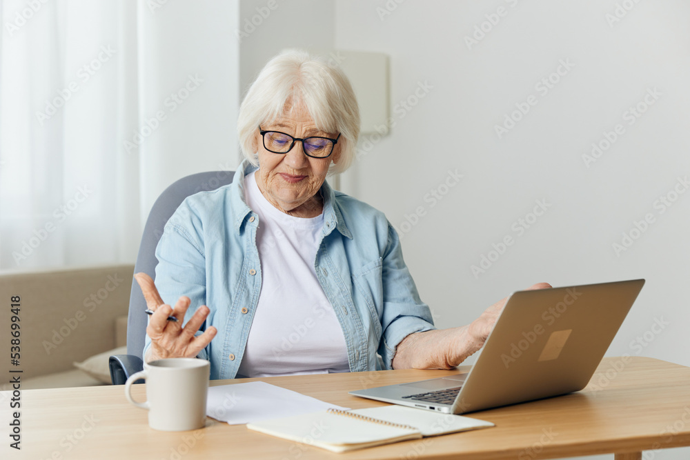 a pleasant, stylish elderly woman is sitting at home in a bright interior and working on a laptop makes notes on a piece of paper. The concept of working from home a pleasant, stylish elderly woman is sitting at home in a bright interior and working on a laptop makes notes on a piece of paper. The concept of working from home