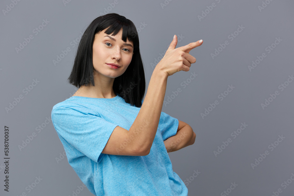 an admiring woman stands on a gray background in a blue T-shirt, smiles a little, points her finger to the side, putting her hand on her hip, turning half sideways an admiring woman stands on a gray background in a blue T-shirt, smiles a little, points her finger to the side, putting her hand on her hip, turning half sideways