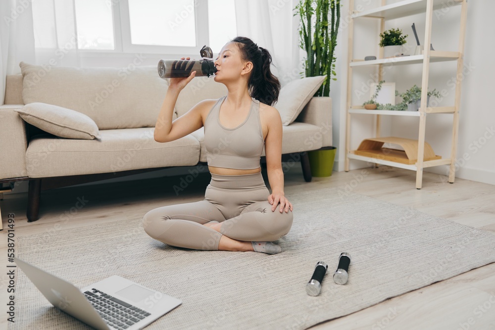 Sporty Asian woman in a yoga suit blogger watching a laptop recording of a sports workout online smiling and drinking water while relaxing at home, lifestyle in sports and good nutrition