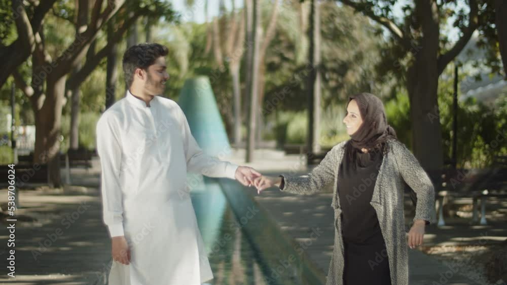 Side view of young muslim couple holding hands in park. Arabic man in white shirt and beautiful woman in hijab looking at each other, talking, having fun on warm summer day. Love, ethnicity concept. Side view of young muslim couple holding hands in park. Arabic man in white shirt and beautiful woman in hijab looking at each other, talking, having fun on warm summer day. Love, ethnicity concept.