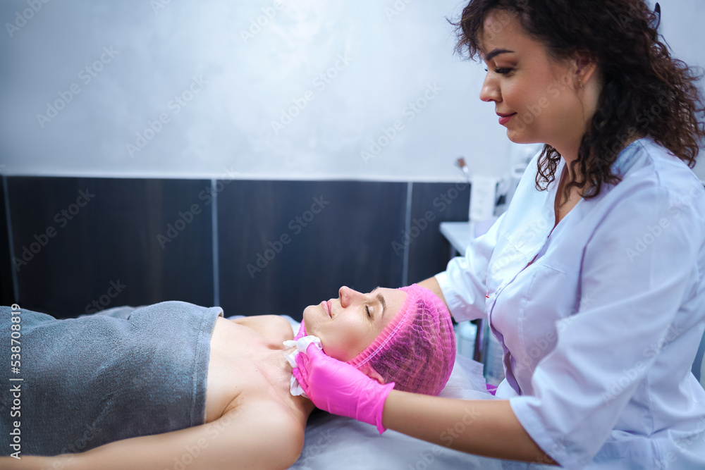 Doctor cosmetologist performing a cosmetic procedure on patient's face in a bright beauty shop and has a fun chatting with her Doctor cosmetologist performing a cosmetic procedure on patient's face in a bright beauty shop and has a fun chatting with her