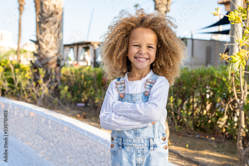 Close up portrait of a smiling child girl in spring park on sunny warm day with arms folded.