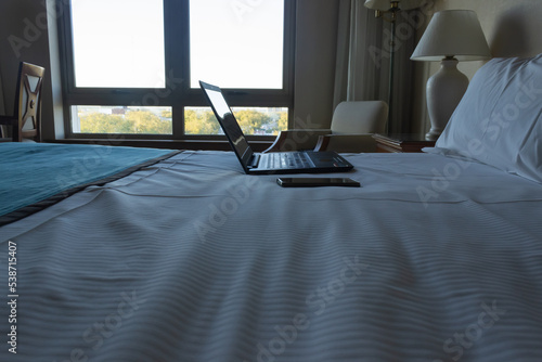 Laptop and cell phone on a bed with pillows and white sheets.