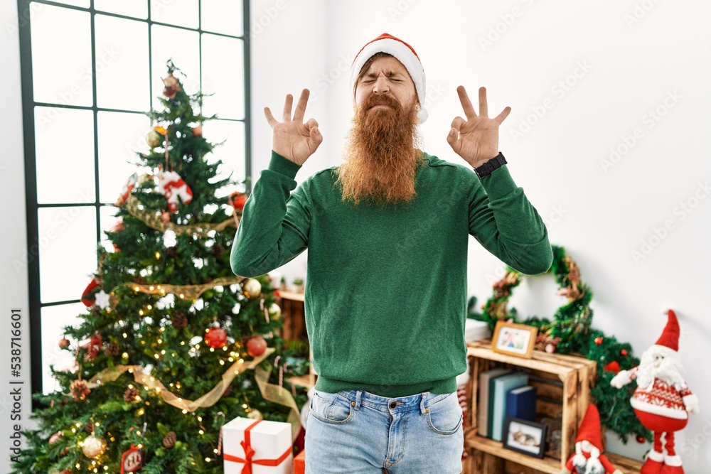 Redhead man with long beard wearing christmas hat by christmas tree relaxed and smiling with eyes closed doing meditation gesture with fingers. yoga concept.