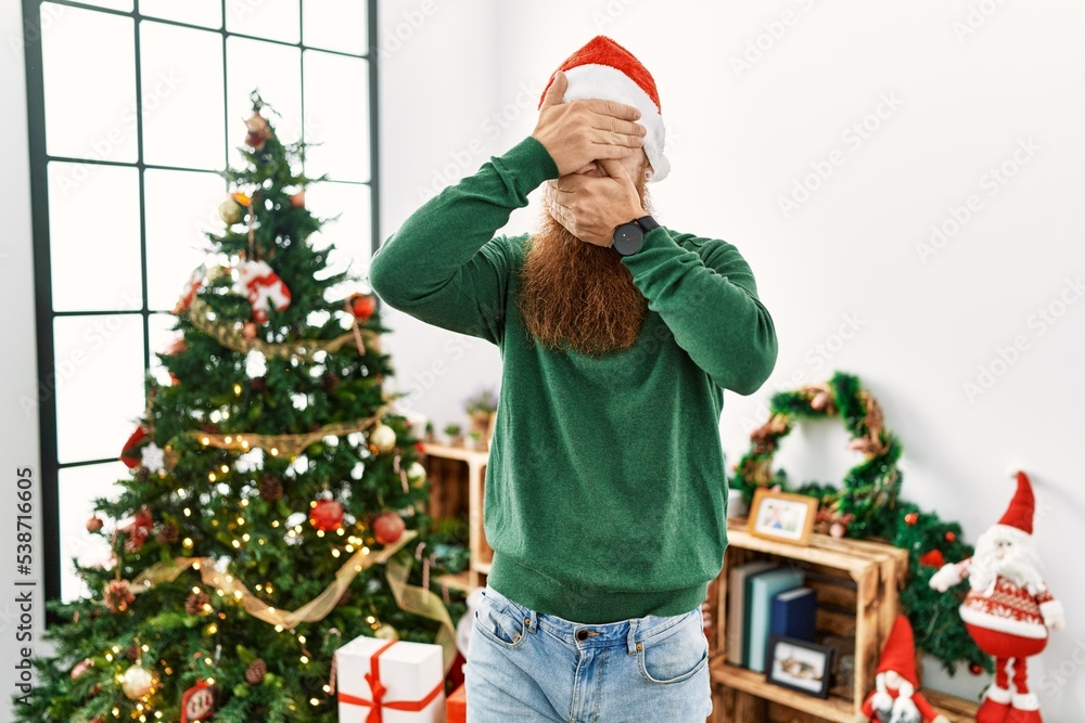 Redhead man with long beard wearing christmas hat by christmas tree covering eyes and mouth with hands, surprised and shocked. hiding emotion Redhead man with long beard wearing christmas hat by christmas tree covering eyes and mouth with hands, surprised and shocked. hiding emotion