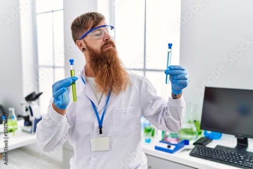 Young redhead man wearing scientist uniform holding test tubes at laboratory