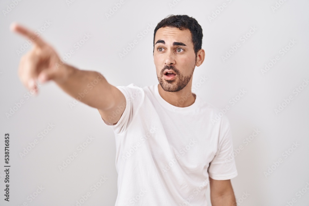 Handsome hispanic man standing over white background pointing with finger surprised ahead, open mouth amazed expression, something on the front Handsome hispanic man standing over white background pointing with finger surprised ahead, open mouth amazed expression, something on the front