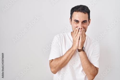 Handsome hispanic man standing over white background laughing and embarrassed giggle covering mouth with hands, gossip and scandal concept