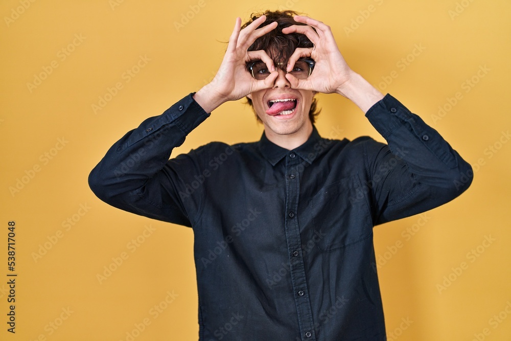 Young man wearing glasses over yellow background doing ok gesture like binoculars sticking tongue out, eyes looking through fingers. crazy expression. Young man wearing glasses over yellow background doing ok gesture like binoculars sticking tongue out, eyes looking through fingers. crazy expression.