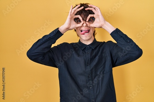 Young man wearing glasses over yellow background doing ok gesture like binoculars sticking tongue out, eyes looking through fingers. crazy expression.