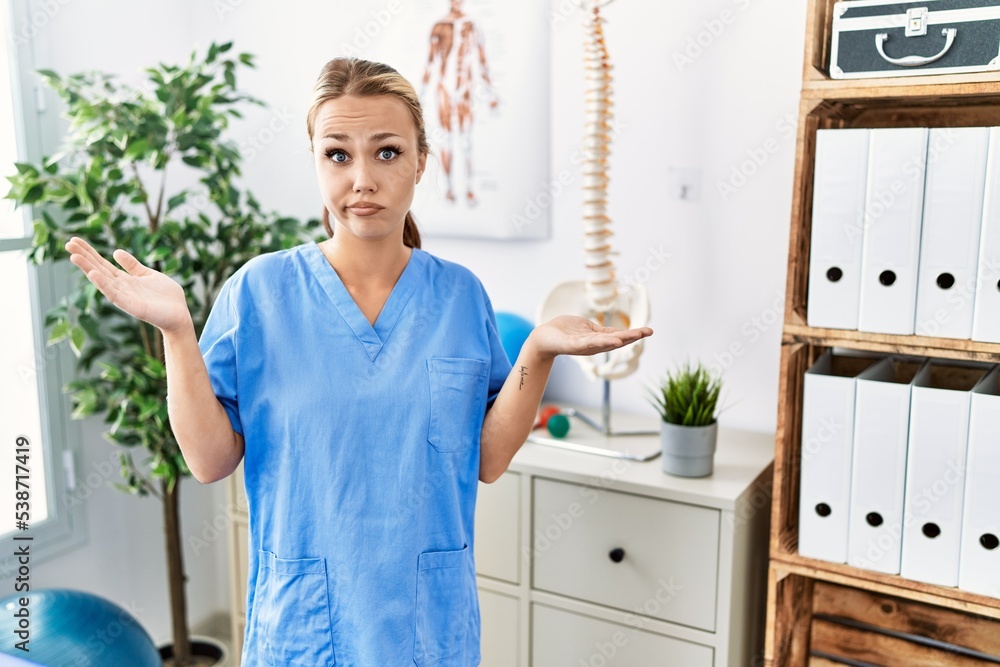 Young caucasian woman working at pain recovery clinic clueless and confused expression with arms and hands raised. doubt concept. Young caucasian woman working at pain recovery clinic clueless and confused expression with arms and hands raised. doubt concept.
