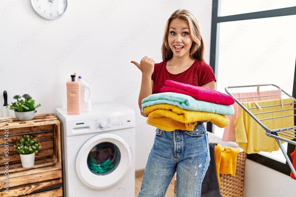 Young caucasian woman holding clean laundry pointing thumb up to the side smiling happy with open mouth Young caucasian woman holding clean laundry pointing thumb up to the side smiling happy with open mouth