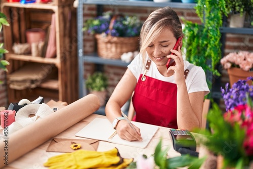 Young blonde woman florist talking on smartphone writing on notebook at florist