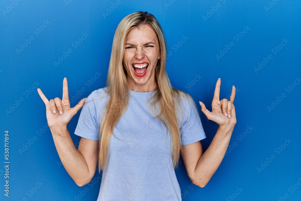 Beautiful blonde woman wearing casual t shirt over blue background shouting with crazy expression doing rock symbol with hands up. music star. heavy concept. Beautiful blonde woman wearing casual t shirt over blue background shouting with crazy expression doing rock symbol with hands up. music star. heavy concept.