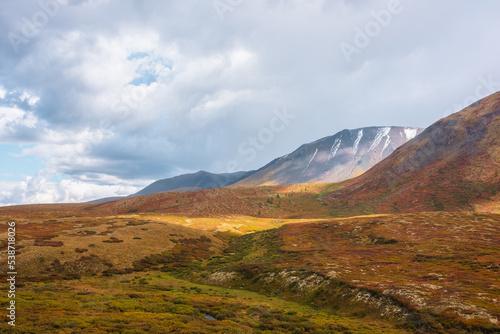 Motley autumn landscape with sunlit hills and mountain range silhouette under dramatic cloudy sky. Vivid autumn colors in mountains. Sunlight on multicolor hills and rainy clouds in changeable weather © Daniil