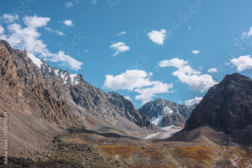 Scenic view to sunlit rocks and large snow mountain top with glacier in autumn sunny day. Vivid autumn colors in high mountains. Motley landscape with sharp rocks and snow mountain peak in bright sun.