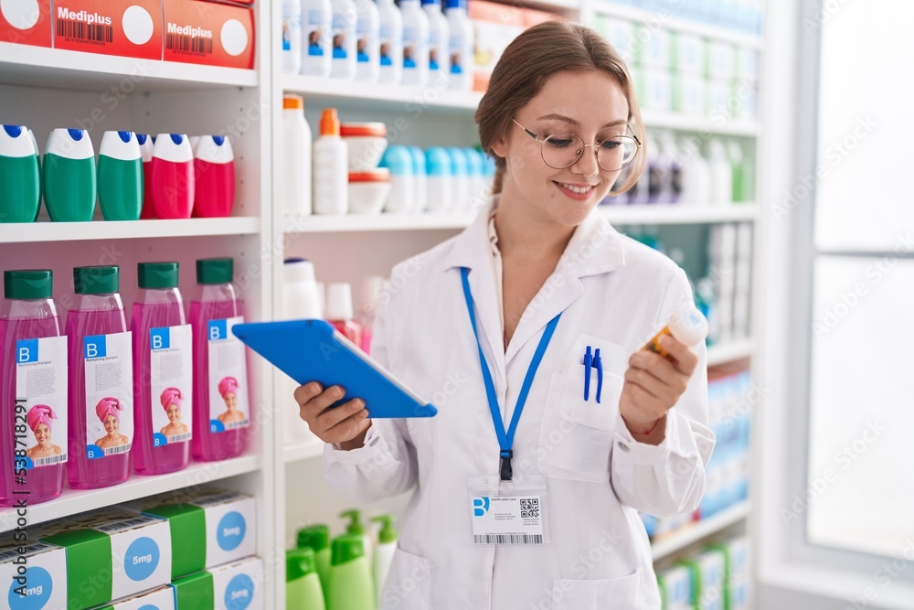 Young blonde woman pharmacist using touchpad holding pills bottle at pharmacy