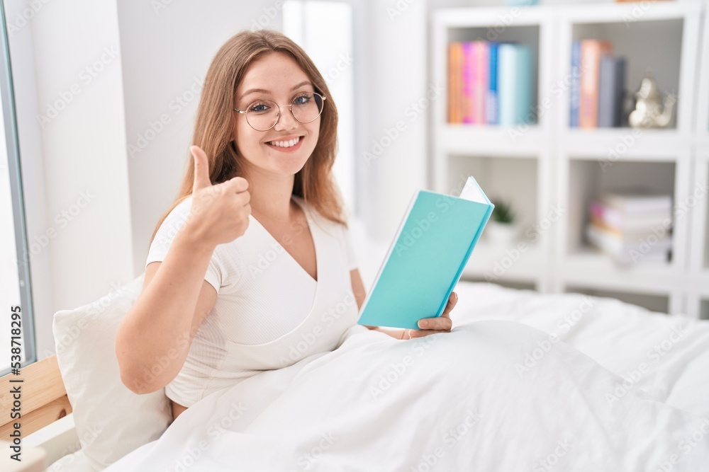 Young caucasian woman sitting on the bed at home reading a book smiling happy and positive, thumb up doing excellent and approval sign Young caucasian woman sitting on the bed at home reading a book smiling happy and positive, thumb up doing excellent and approval sign