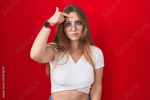 Young caucasian woman standing over red background pointing unhappy to pimple on forehead, ugly infection of blackhead. acne and skin problem