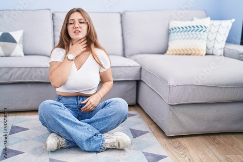 Young caucasian woman sitting on the floor at the living room touching painful neck, sore throat for flu, clod and infection