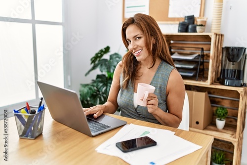 Young latin woman business worker using laptop and drinking coffee at office