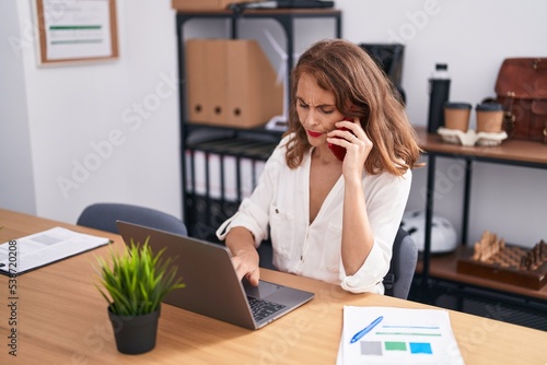 Young beautiful hispanic woman business worker talking on smartphone using laptop at office