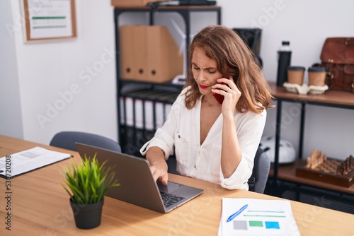 Young beautiful hispanic woman business worker talking on smartphone using laptop at office