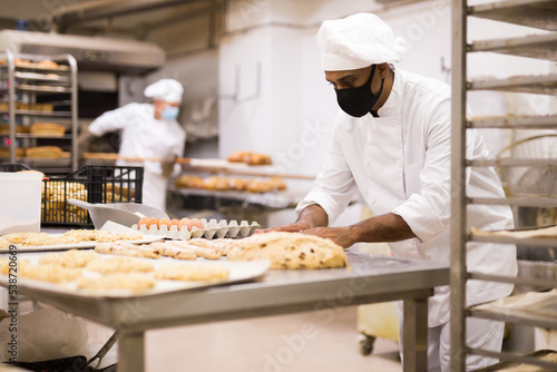 Working at bakery during coronavirus outbreak, man in mask for viral protection kneading dough and shaping baguettes on steel countertop in industrial kitchen