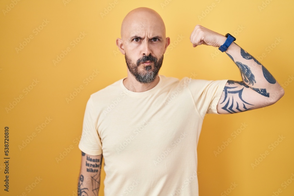 Hispanic man with tattoos standing over yellow background strong person showing arm muscle, confident and proud of power Hispanic man with tattoos standing over yellow background strong person showing arm muscle, confident and proud of power