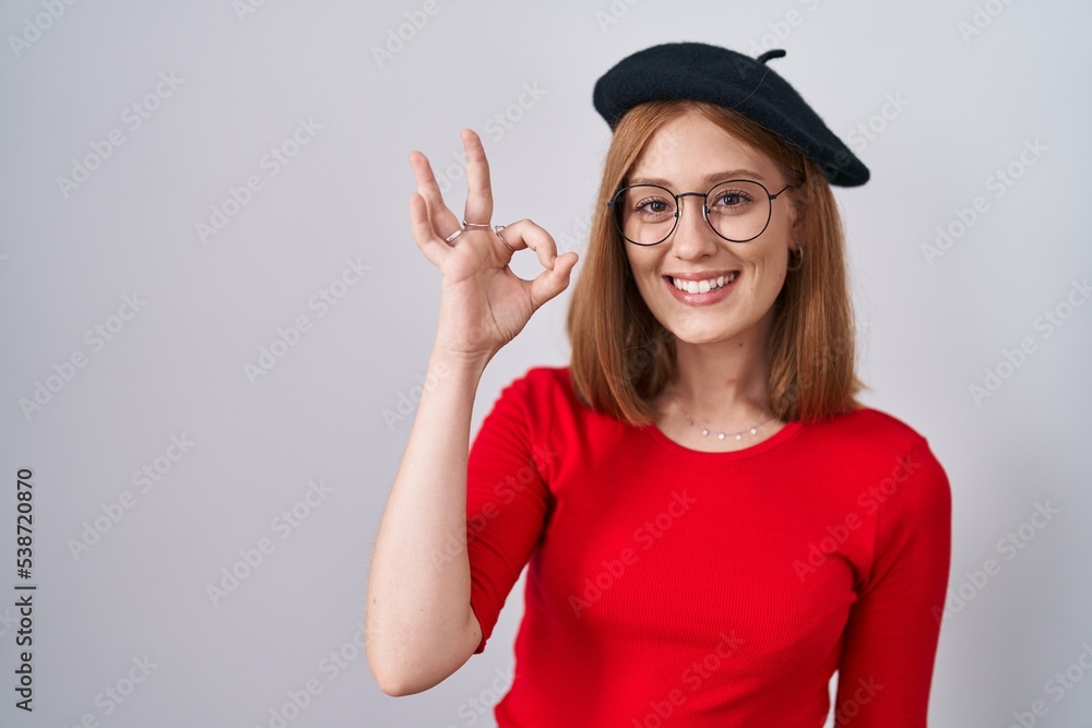 Young redhead woman standing wearing glasses and beret smiling positive doing ok sign with hand and fingers. successful expression. Young redhead woman standing wearing glasses and beret smiling positive doing ok sign with hand and fingers. successful expression.