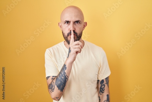 Hispanic man with tattoos standing over yellow background asking to be quiet with finger on lips. silence and secret concept. © Krakenimages.com