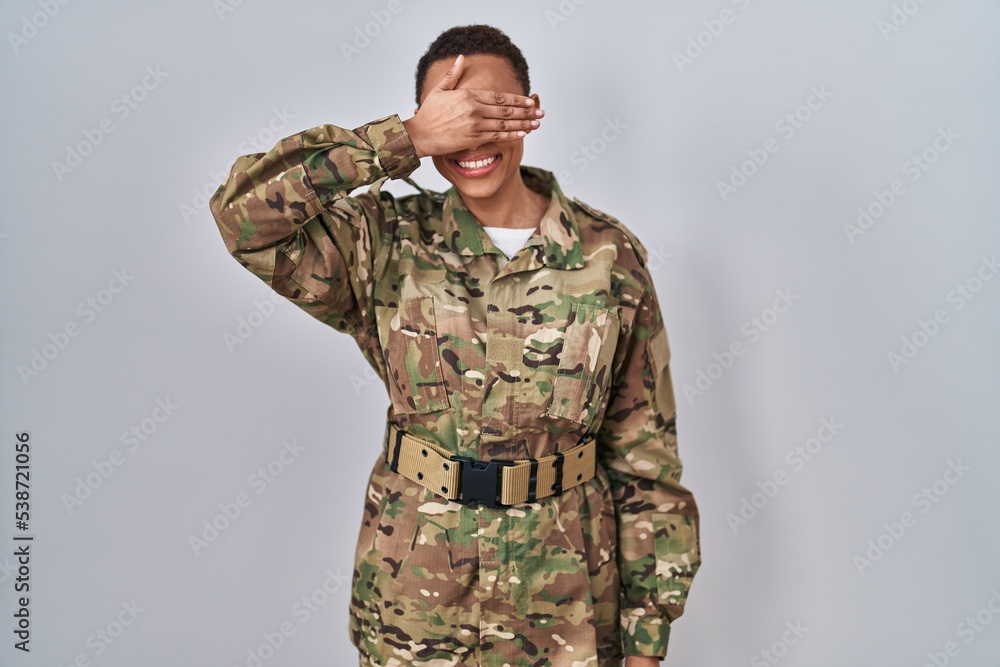 Beautiful african american woman wearing camouflage army uniform smiling and laughing with hand on face covering eyes for surprise. blind concept.