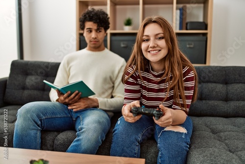 Woman playing video game while unhappy boyfriend read book at home.