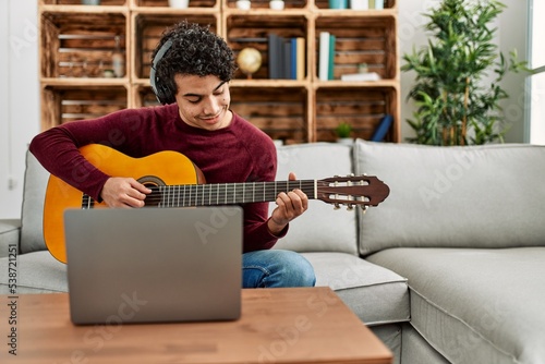 Young hispanic man having online classical guitar lesson sitting on the sofa at home.