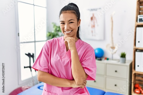 Young hispanic woman working at pain recovery clinic looking confident at the camera smiling with crossed arms and hand raised on chin. thinking positive.